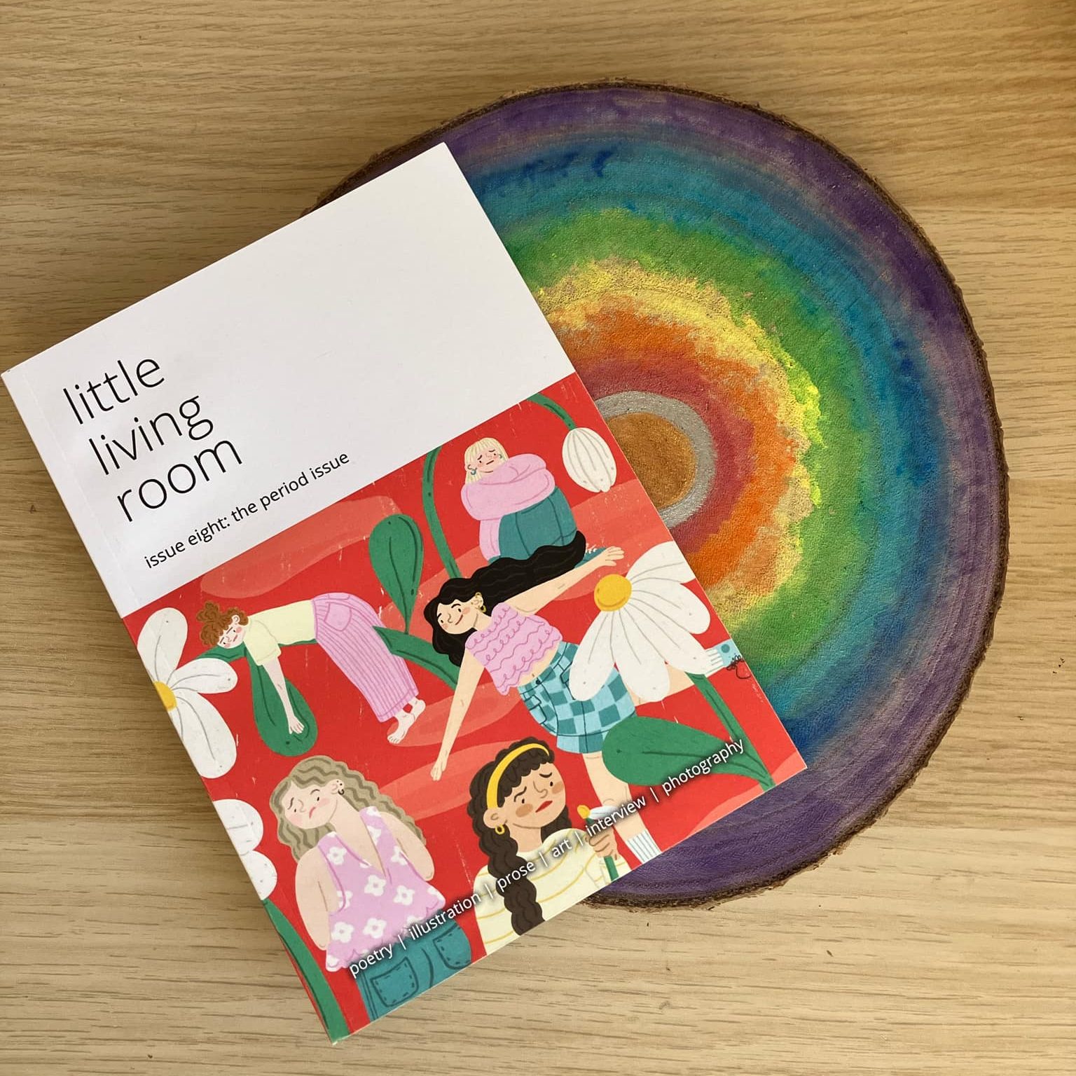 A colorful magazine titled 'little living room, issue eight: the period issue' featuring illustrations of diverse women surrounded by flowers, resting on a rainbow-colored wooden slice on a light wooden surface.