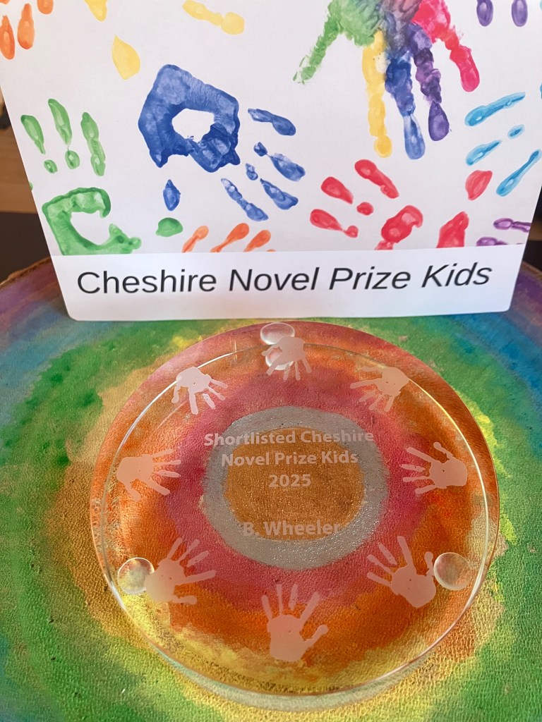 Circular glass award with handprints etched arounds it reading "Shortlisted Cheshire Novel Prize Kids 2025 B Wheeler".  A postcard titled "Cheshire Novel Prize Kids" with colourful painted hand prints is aside it. 